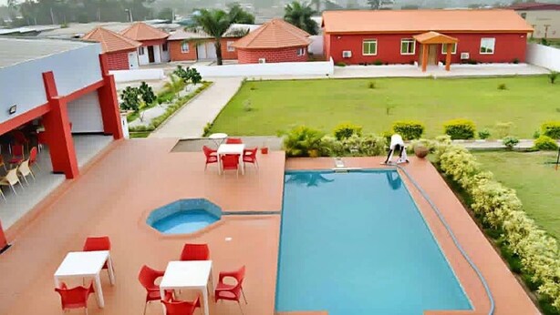 Une piscine entourée d’un bâtiment rouge et d’une pelouse, avec des chaises et des tables rouges.