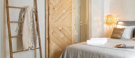 Un dormitorio con un armario de madera, una escalera, una cama con colcha gris, dos cestas tejidas y una lámpara fijada a la pared.