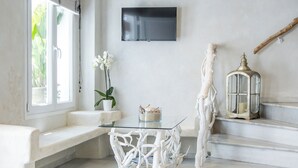 A modern living room with a white bench, a glass table, a potted orchid, a white sculpture, a lantern, and a flat-screen TV on the wall. - Terra Fos Naxos Studios (Naxos)