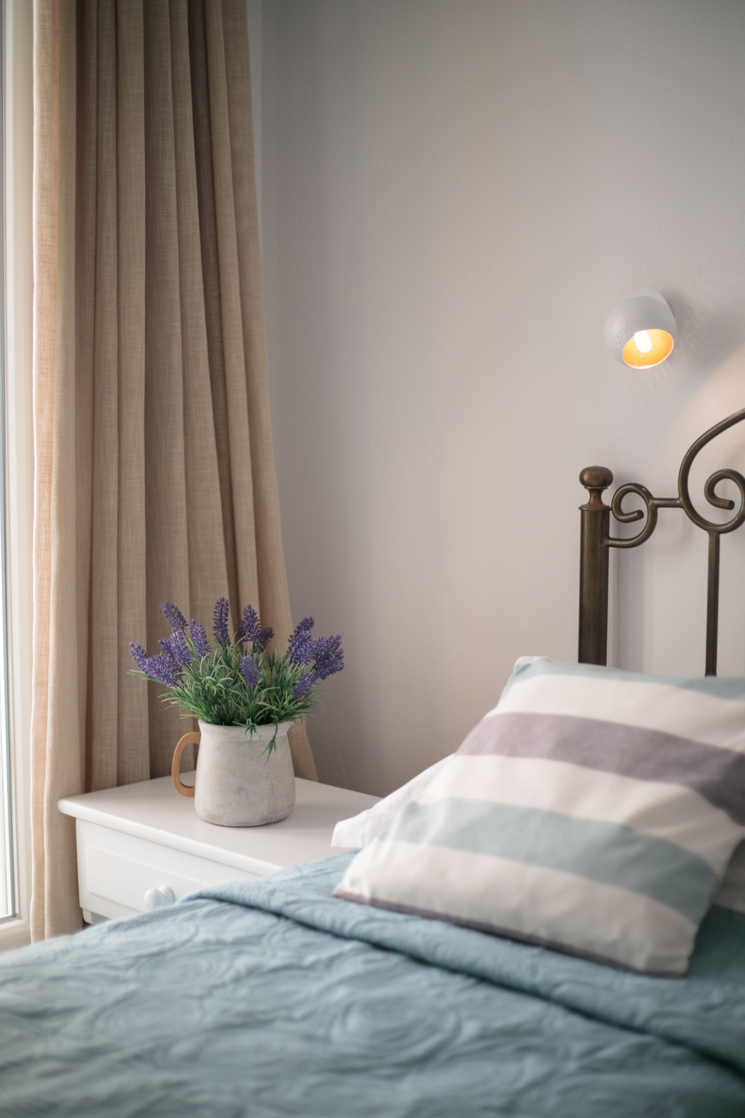 Un dormitorio con cama, mesita de noche con planta de lavanda en maceta, lámpara de pared y ventana con cortinas.