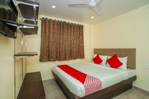 Une chambre d’hôtel avec un lit, un ventilateur de plafond, une fenêtre avec des rideaux à motifs et une petite étagère.