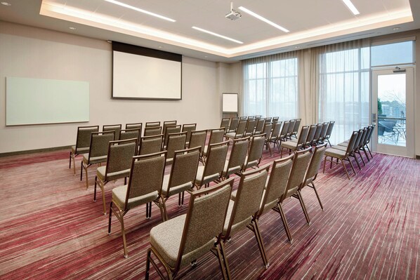 Un salón de conferencias con filas de sillas, una pantalla de proyector y un ventanal grande.