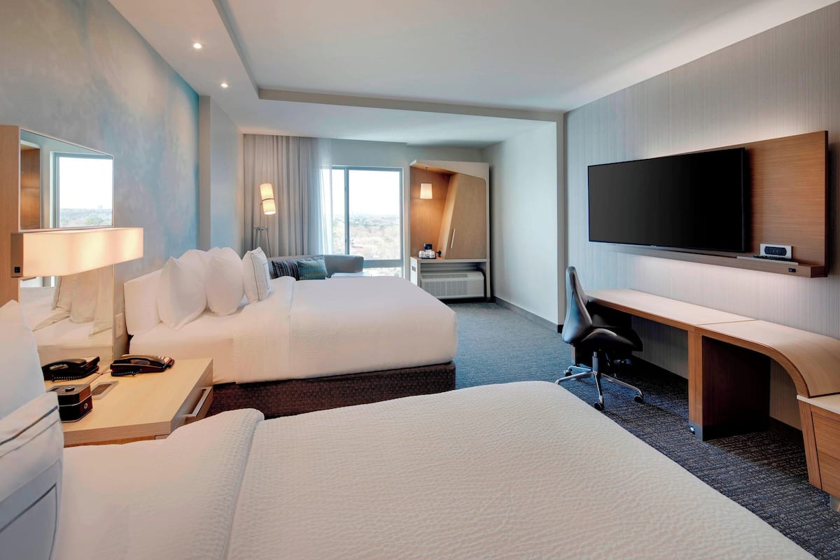 Ein Hotelzimmer mit zwei Betten, einem Schreibtisch, einem Flachbildfernseher, einer Lampe und einem großen Fenster.