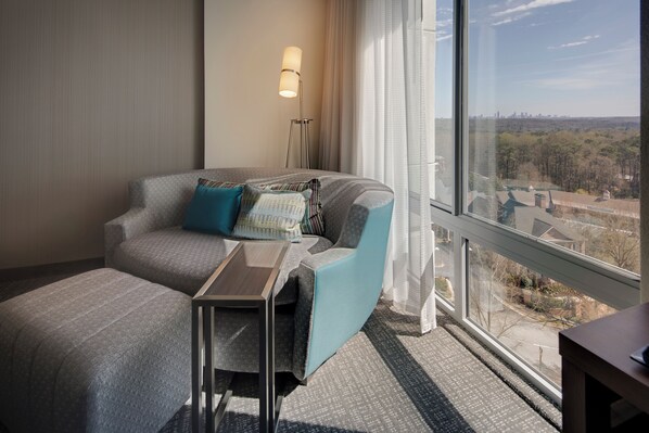A hotel room with a large window, a sofa, a small table, and a lamp.