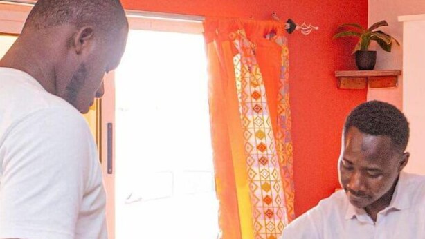 Two individuals in a room with a red wall and patterned curtains.