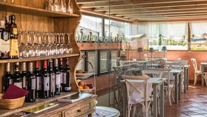 Un restaurante con una decoración rústica, que presenta estantes de madera con botellas y copas de vino, un área de comedor con sillas blancas y vistas a la vegetación a través de amplias ventanas.