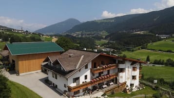 A two-story building with a balcony, surrounded by green fields and mountains.