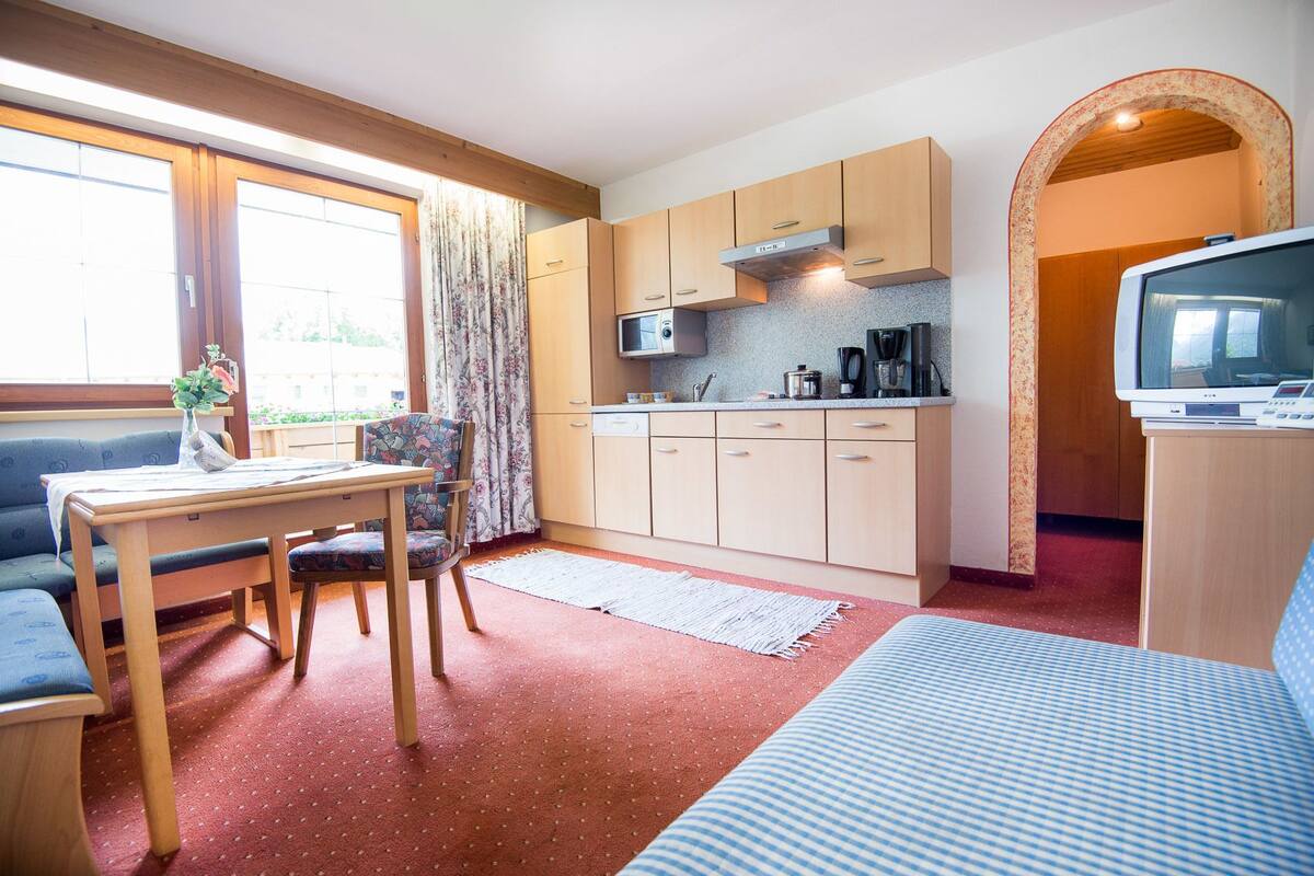 A compact kitchen with wooden cabinets, a microwave, and a coffee maker. There is a dining table with chairs, a sofa, and a TV on a stand.
