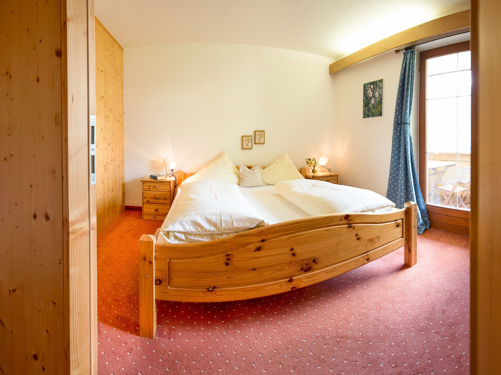 A wooden bed in a room with a red carpet, a wooden wardrobe, and a window with a curtain.