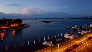 A marina at dusk with boats docked, a road leading to the water, and a building with lights on the left. - ApartHotel Capitol (Grado)