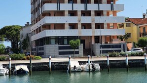 A multi-story building with balconies, a dock with boats, and a clear sky. - ApartHotel Capitol (Grado)