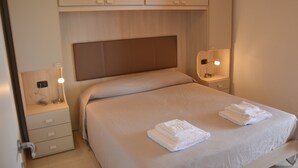 A neatly made bed with a headboard, a bedside table with a lamp, and a drawer unit. - ApartHotel Capitol (Grado)