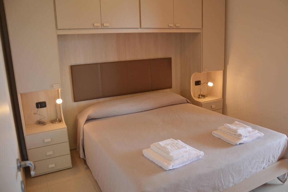 A neatly made bed with a headboard, a bedside table with a lamp, and a drawer unit.