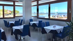 A restaurant with tables and chairs, large windows with a view of a body of water and buildings. - ApartHotel Capitol (Grado)