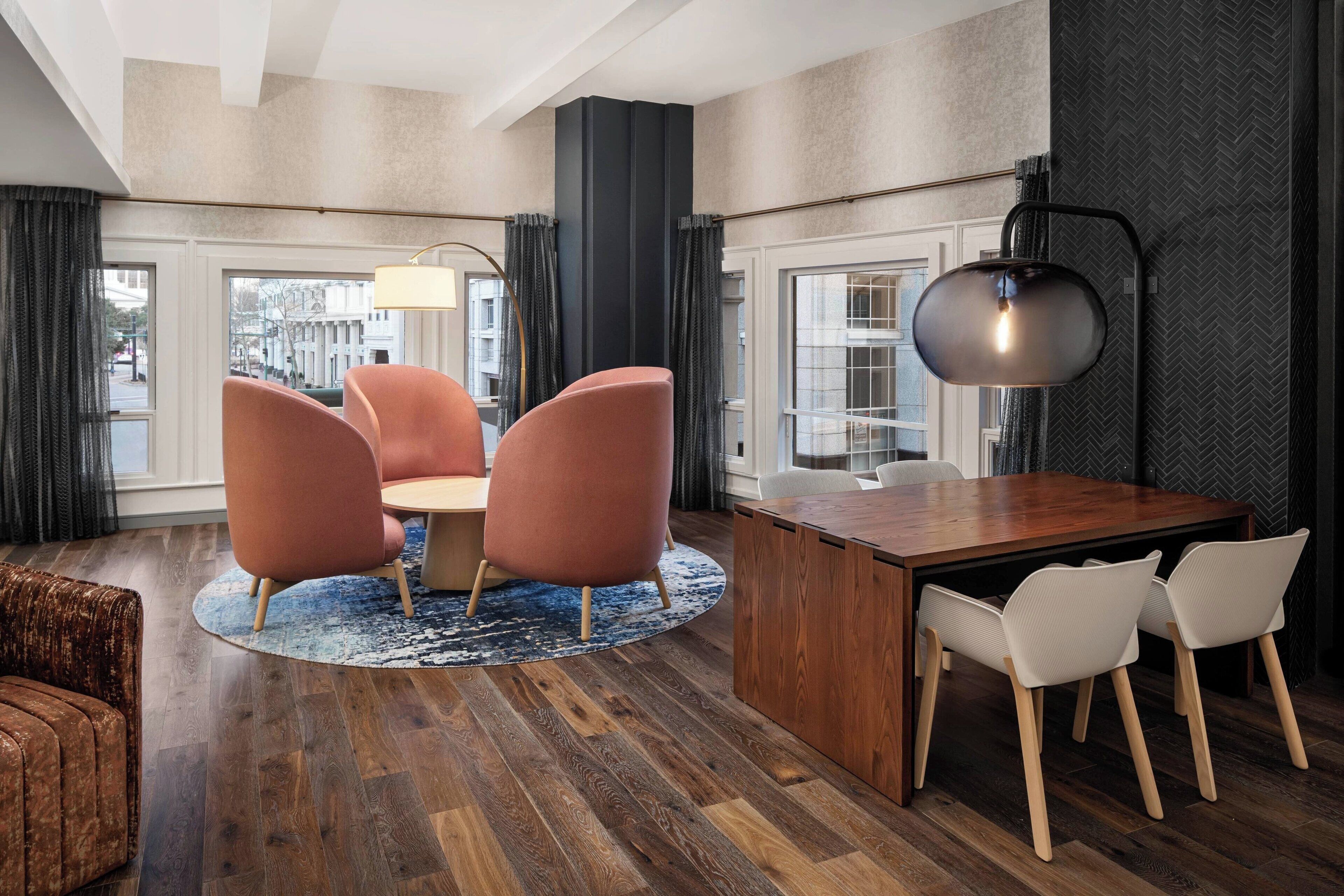 Una habitación de hotel moderna con una mesa de comedor de madera, dos sillas rosas, una alfombra redonda y un amplio ventanal con cortinas.