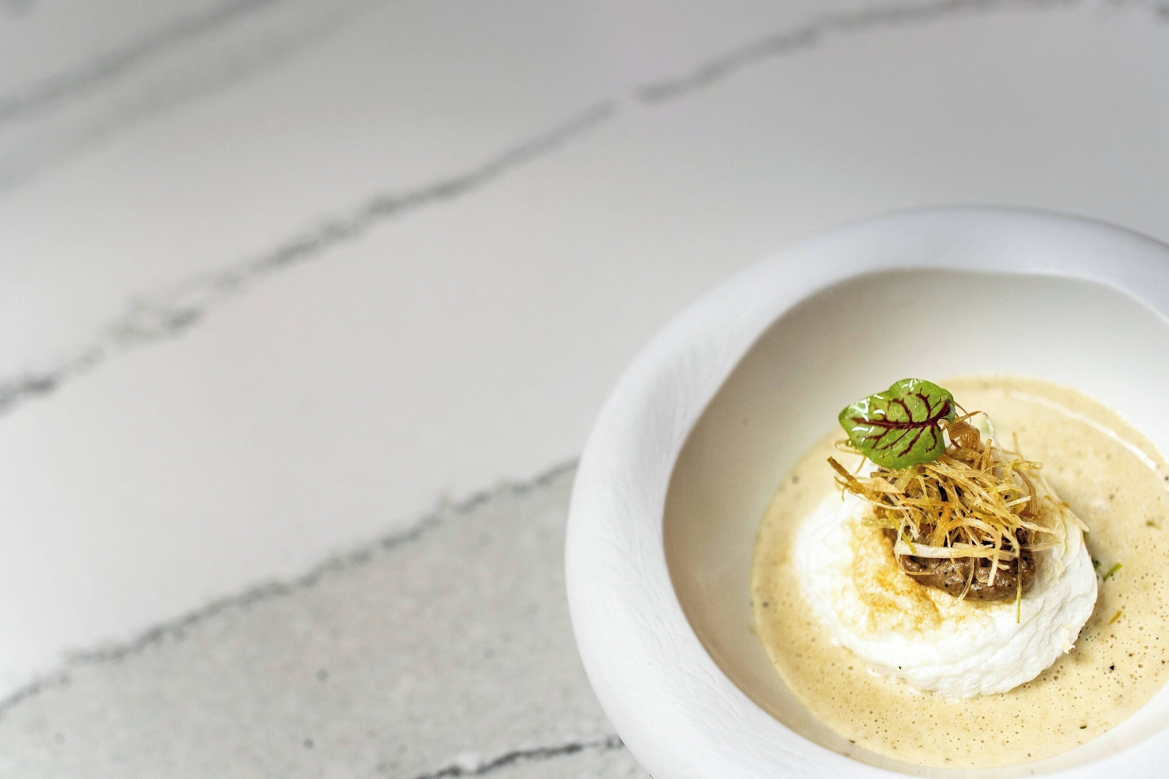 Un tazón blanco con una crema para sopa y una porción de alimento blanco, posiblemente un postre, sobre una superficie de mármol.