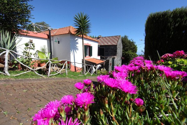 A white building with a red-tiled roof, surrounded by lush greenery and vibrant pink flowers.