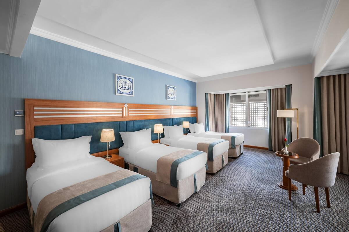 A hotel room with three beds, a sitting area with a chair, a table, and a lamp.