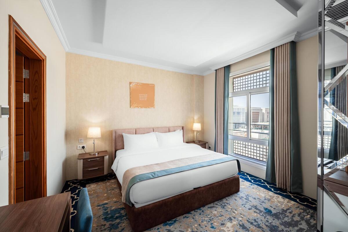 A hotel room with a large bed, a wooden wardrobe, a nightstand with a lamp, a window with a view of the city, and a blue patterned carpet.