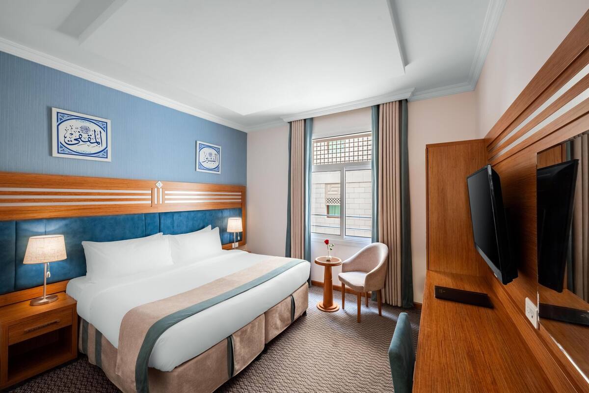 A hotel room with a large bed, a TV, a small table with a chair, and two wall-mounted picture frames.