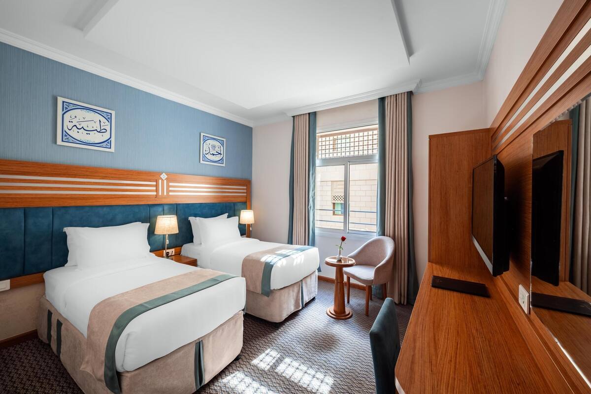 A hotel room with two beds, a TV, a small table with a chair, and a window with curtains.