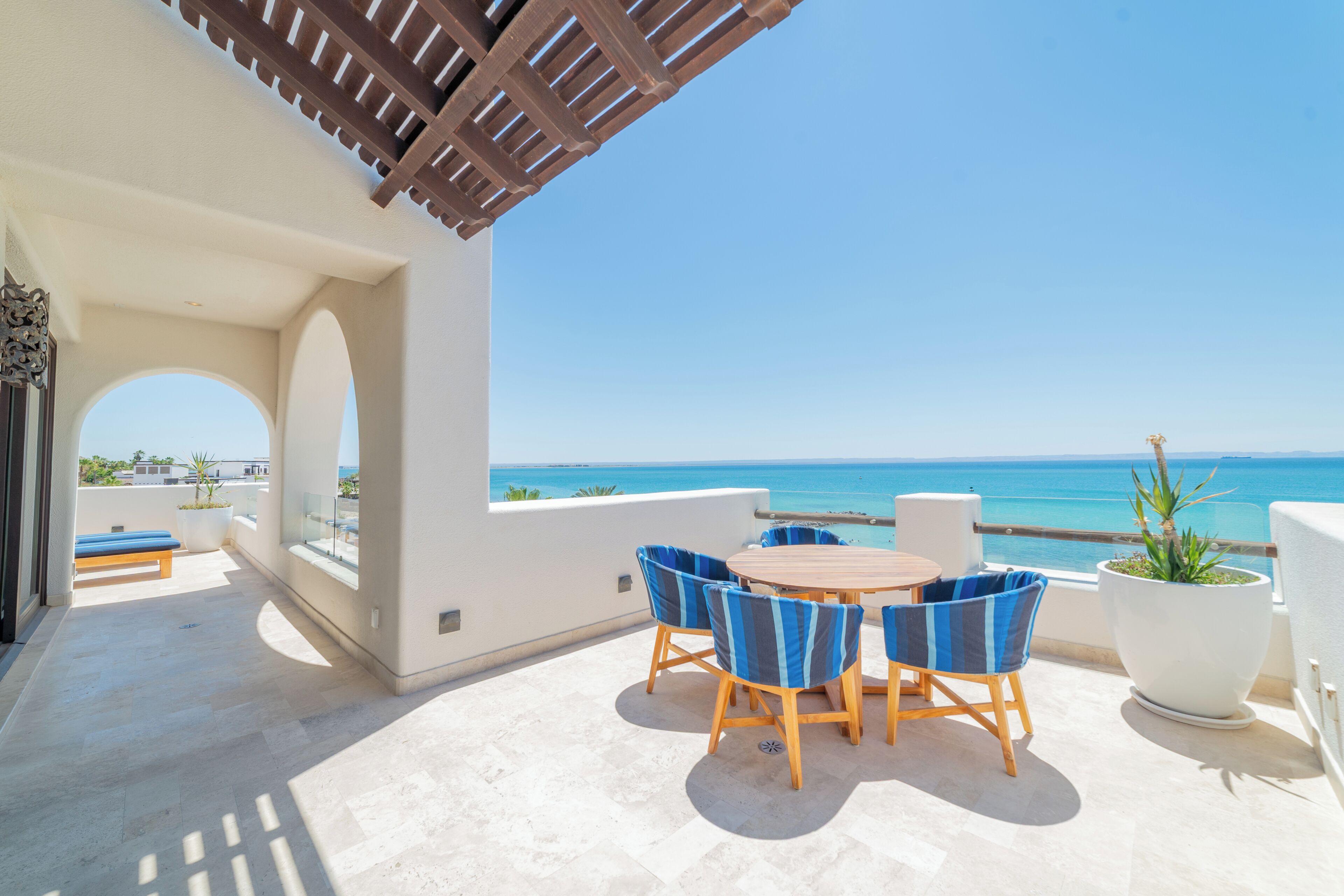 A terrace with a view of the sea, a round table, and blue chairs.