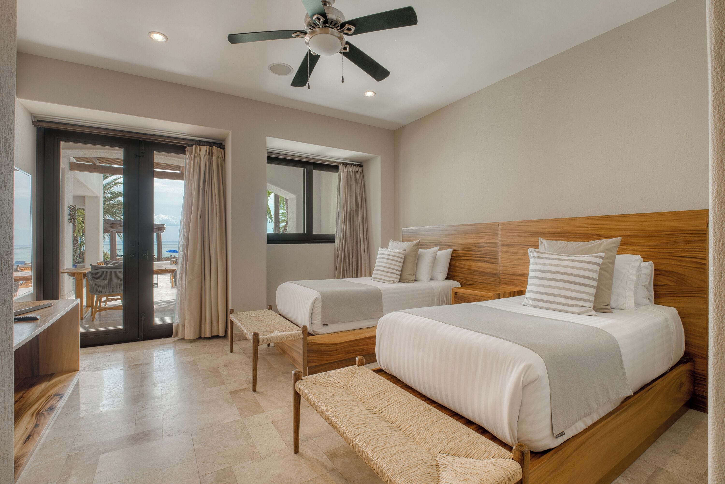 A hotel room with two beds, a ceiling fan, a bench, a desk, and a view of the outdoors.