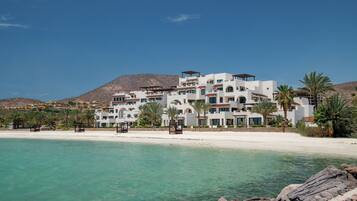 Un hotel frente a la playa con varios edificios blancos, una playa de arena y agua turquesa cristalina.