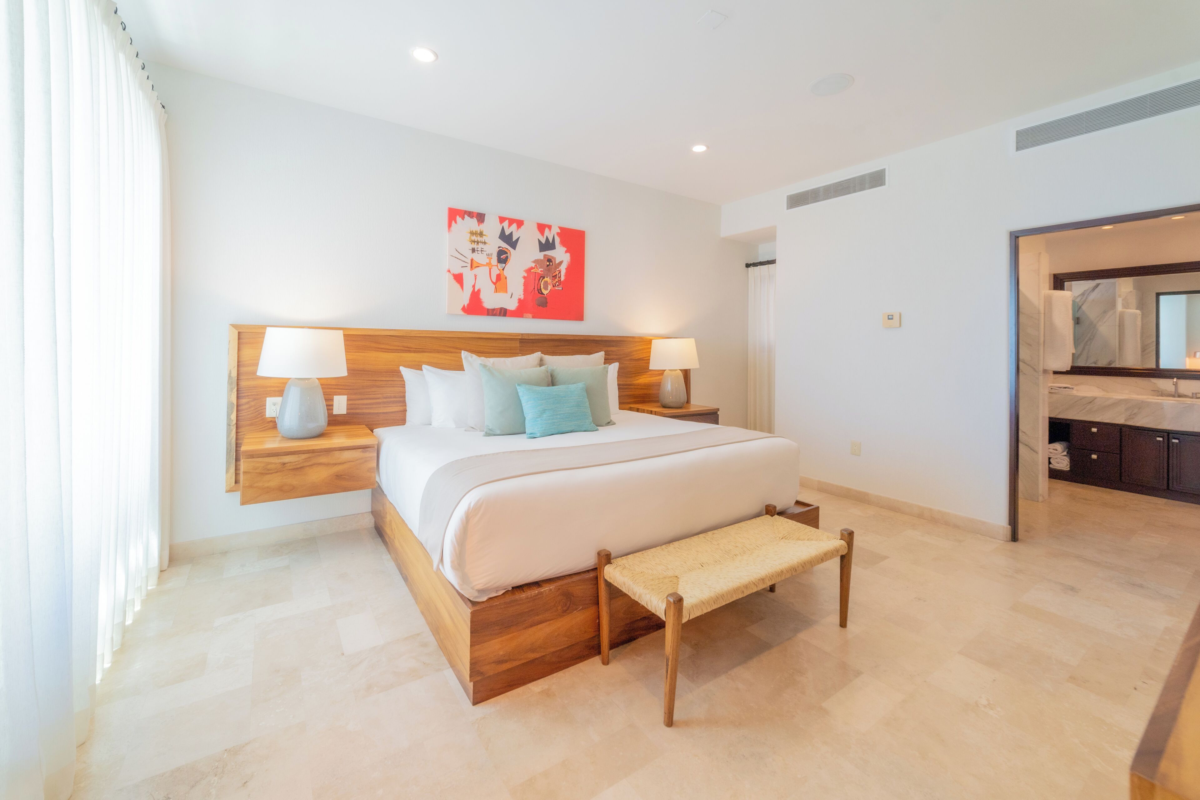 A modern hotel room with a large bed, bedside lamps, a wooden headboard, a bench, and a bathroom visible through an open door.