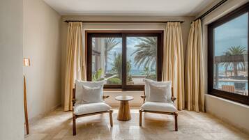 A room with two white cushioned chairs facing a large window with curtains, a round wooden table, and a view of palm trees and the sea.