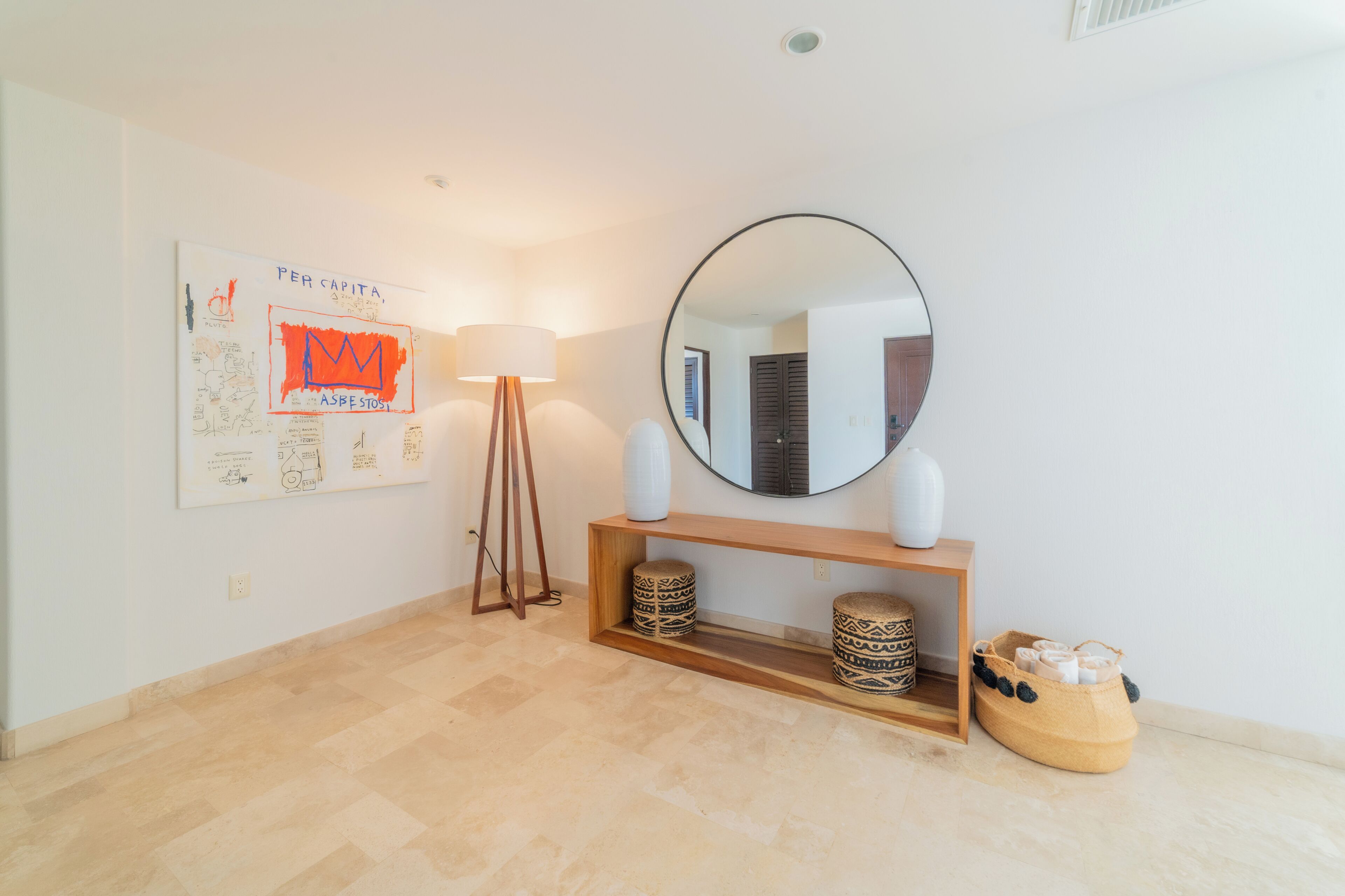 Une pièce comprenant une console en bois, un miroir rond, une lampe, des tabourets à motifs et un mur orné d’une affiche.