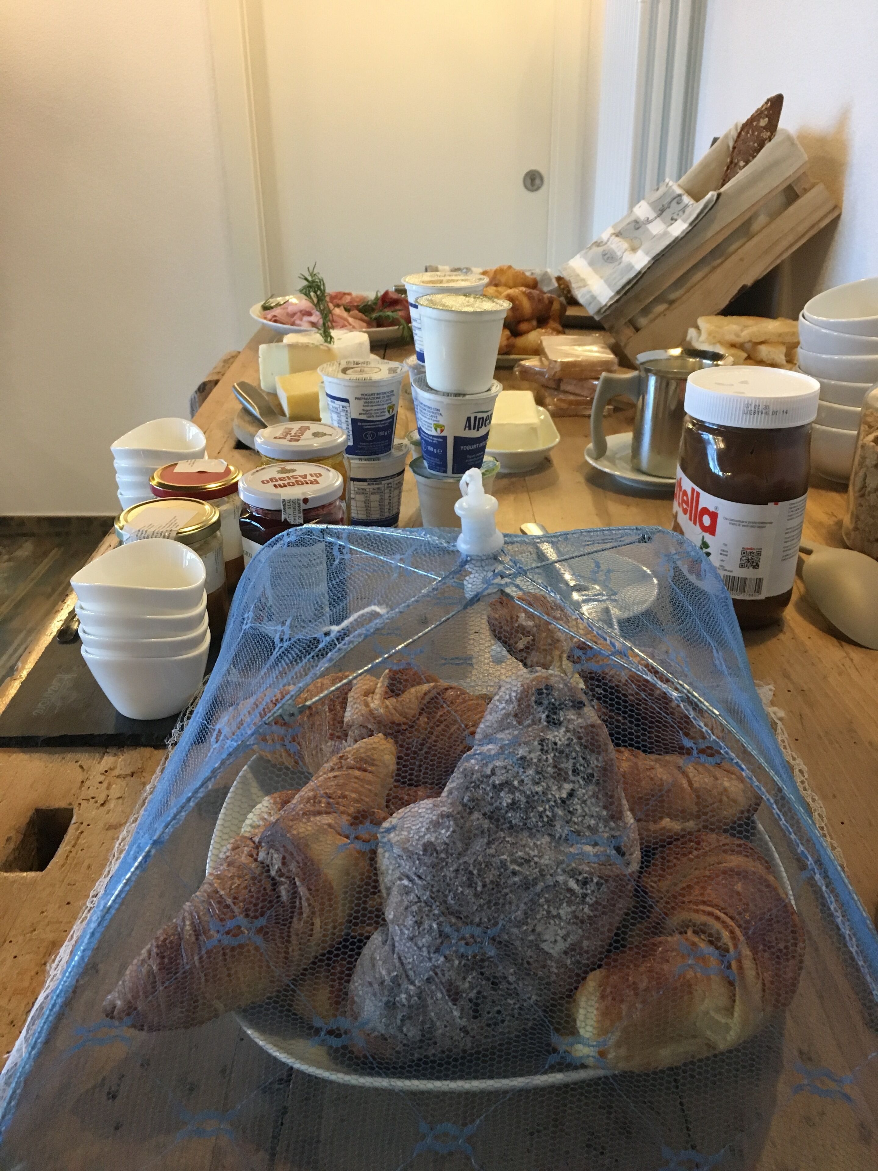 Uma mesa com diversos alimentos, incluindo potes de geleia, uma garrafa de Nutella e uma cesta de croissants.