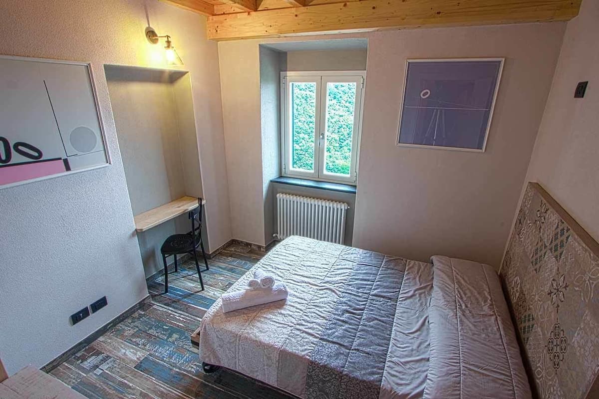 A bedroom with a bed, a desk, a chair, a radiator, and a window with a view of greenery.
