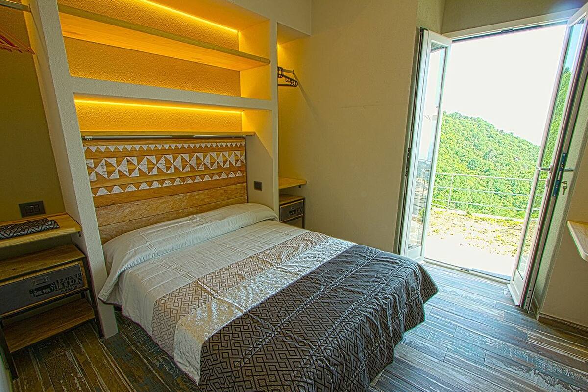 A hotel room with a bed, a bedside table, a nightstand, and a view of a green landscape through a glass door.