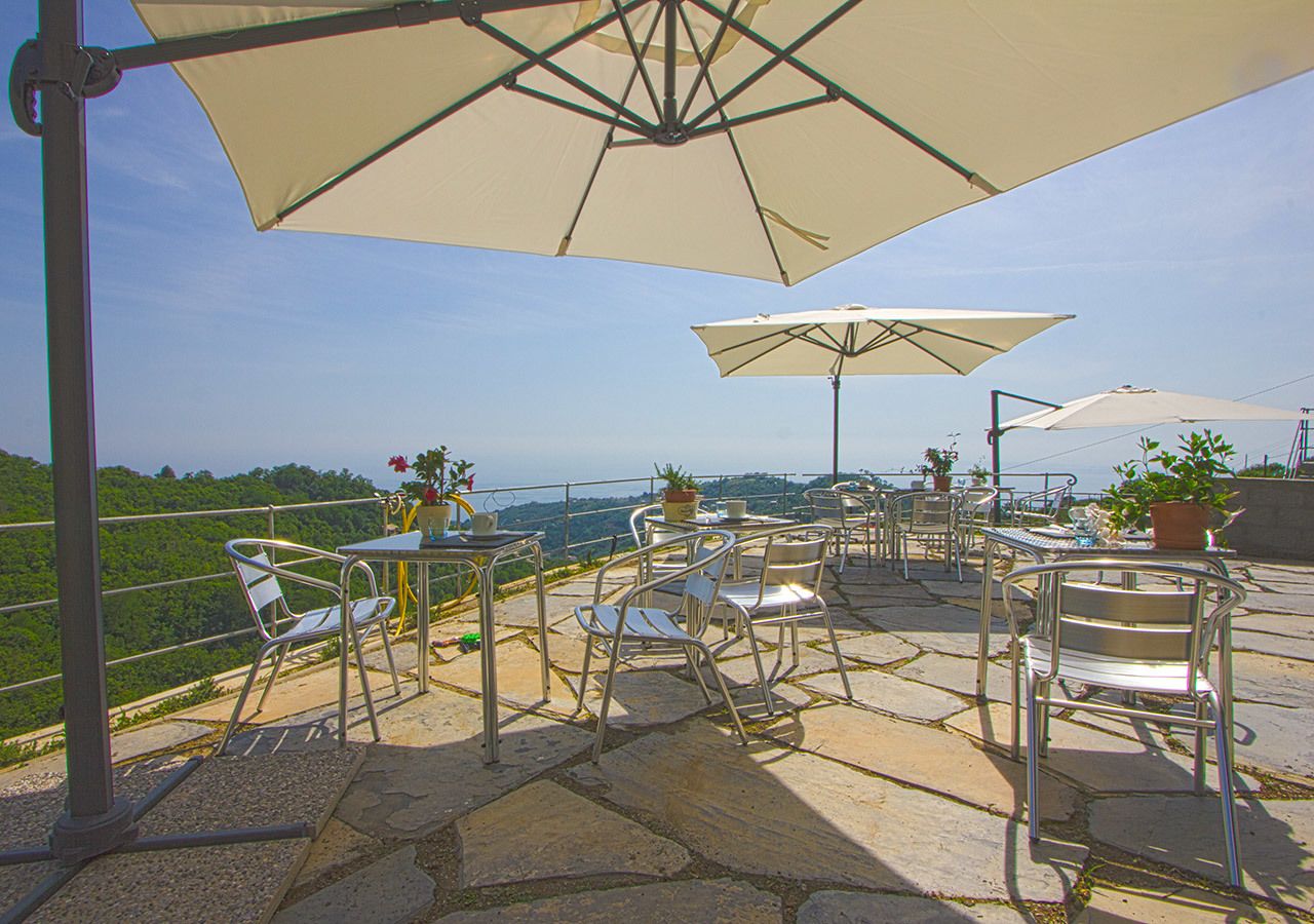 An outdoor patio with stone flooring, metal chairs, and tables, featuring umbrellas and potted plants.