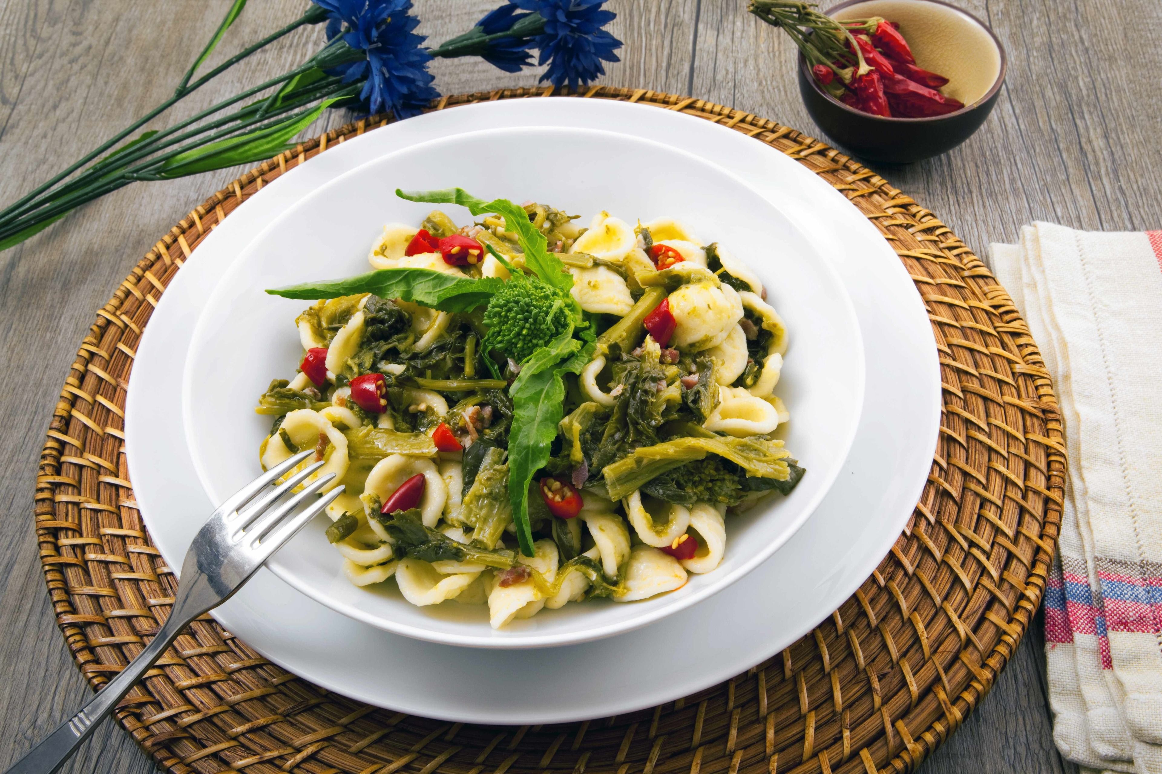 Un plato de pasta con verduras y hierbas sobre una fuente blanca, colocado sobre un tapete tejido.