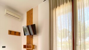Habitación de hotel con cama, televisor montado, paneles de madera y un ventanal con cortinas traslúcidas.