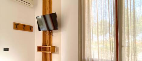 A hotel room with a bed, a mounted TV, wooden paneling, and a large window with sheer curtains.