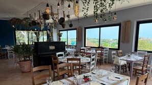 Un restaurante con varias mesas dispuestas para comensales, amplias ventanas que ofrecen vistas panorámicas y plantas y luces colgantes que crean un ambiente cálido.