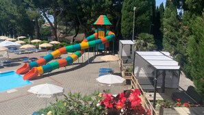 Un área de piscina con un tobogán acuático colorido, sillas de descanso, sombrillas y un pequeño edificio con techo rojo.