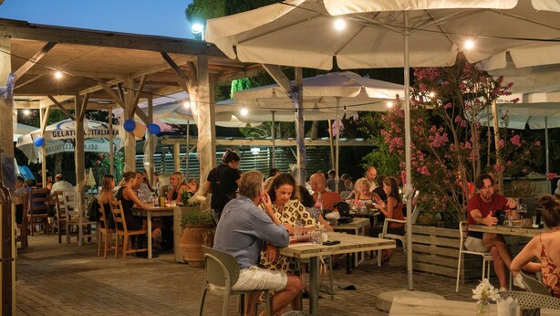 Un área de comedor al aire libre con mesas y sillas bajo sombrillas, rodeada de plantas en macetas y un letrero para Gelateria Italiana.