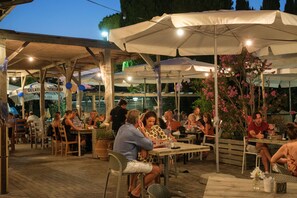 Un espace de restauration en plein air avec des tables et des chaises sous des parasols, entouré de plantes en pot et d’une enseigne indiquant « Gelateria Italiana ».