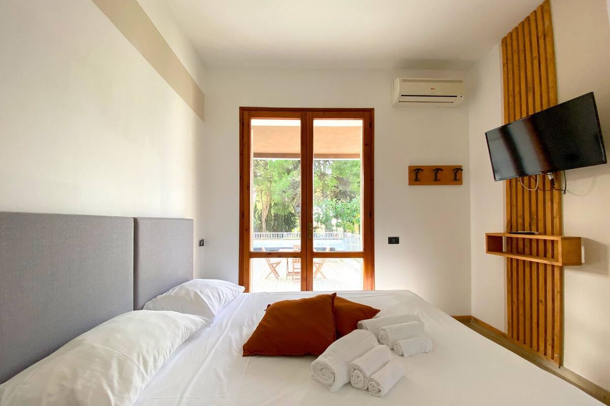 A hotel room with a bed, white linens, a TV mounted on the wall, and a view of the outdoors through a door.