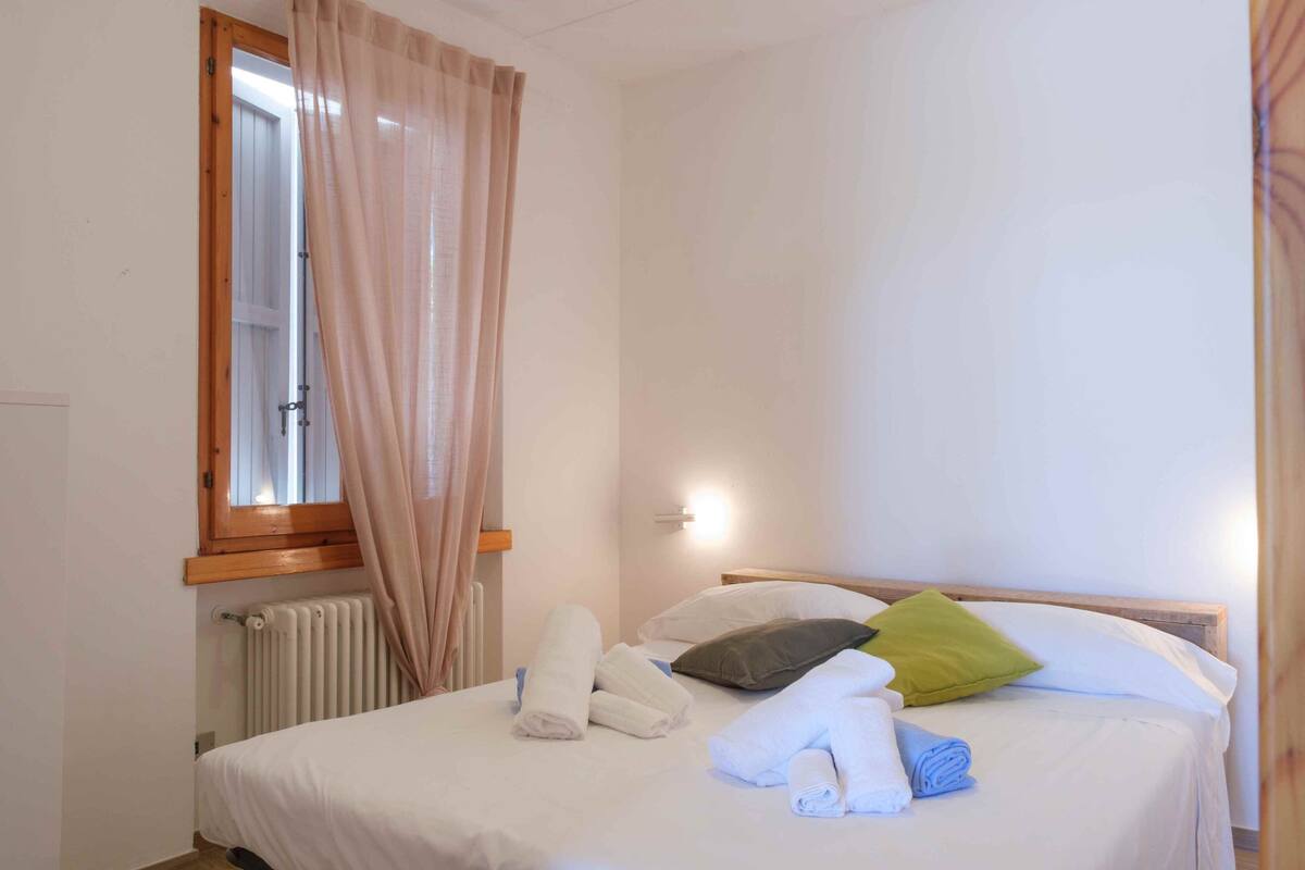 A neatly made bed with rolled towels, a window with curtains, and a wall-mounted light fixture.