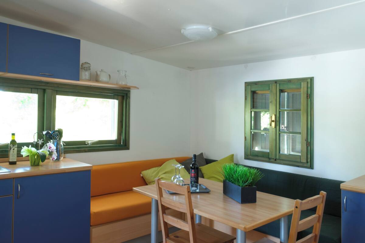 A compact kitchen with a wooden table, a green cushioned bench, and a window with green shutters.