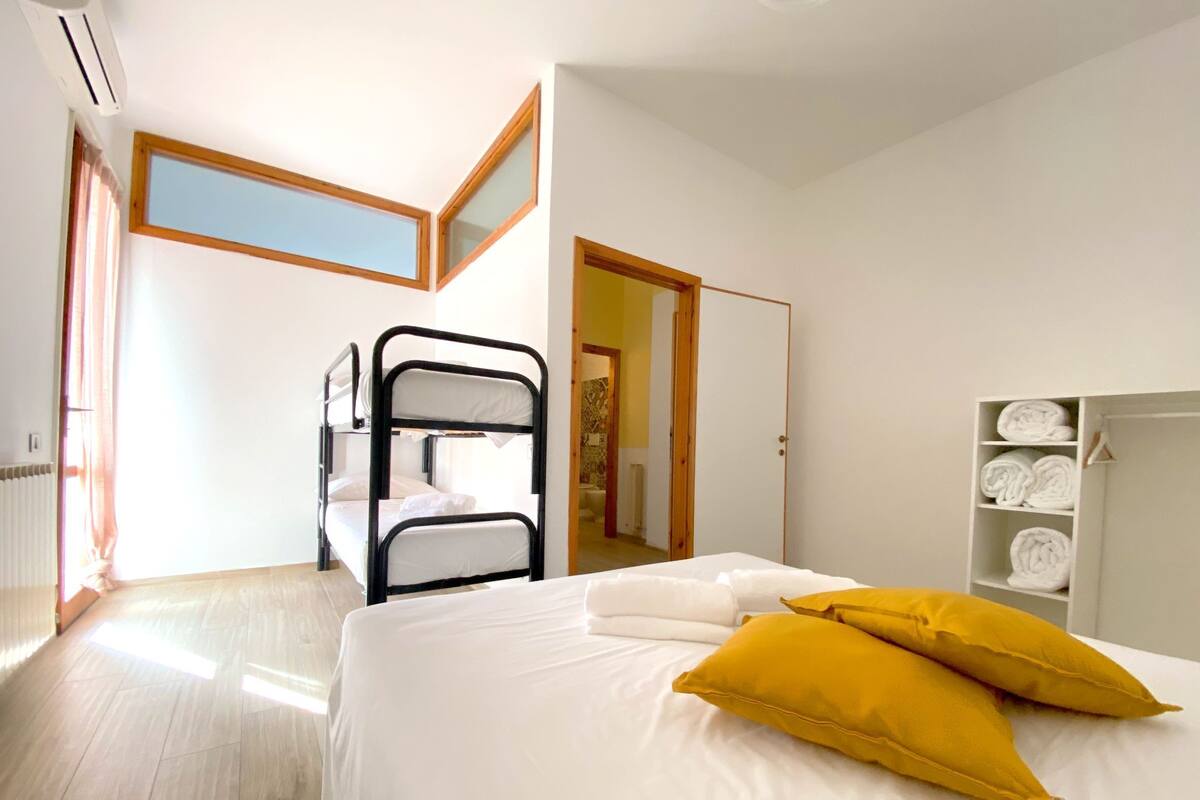 A room with a bunk bed, a bed with yellow pillows, a wardrobe with towels, and a window with curtains.