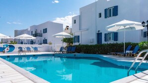 Una piscina con sillas de descanso y sombrillas, rodeada de edificios blancos con contraventanas azules.