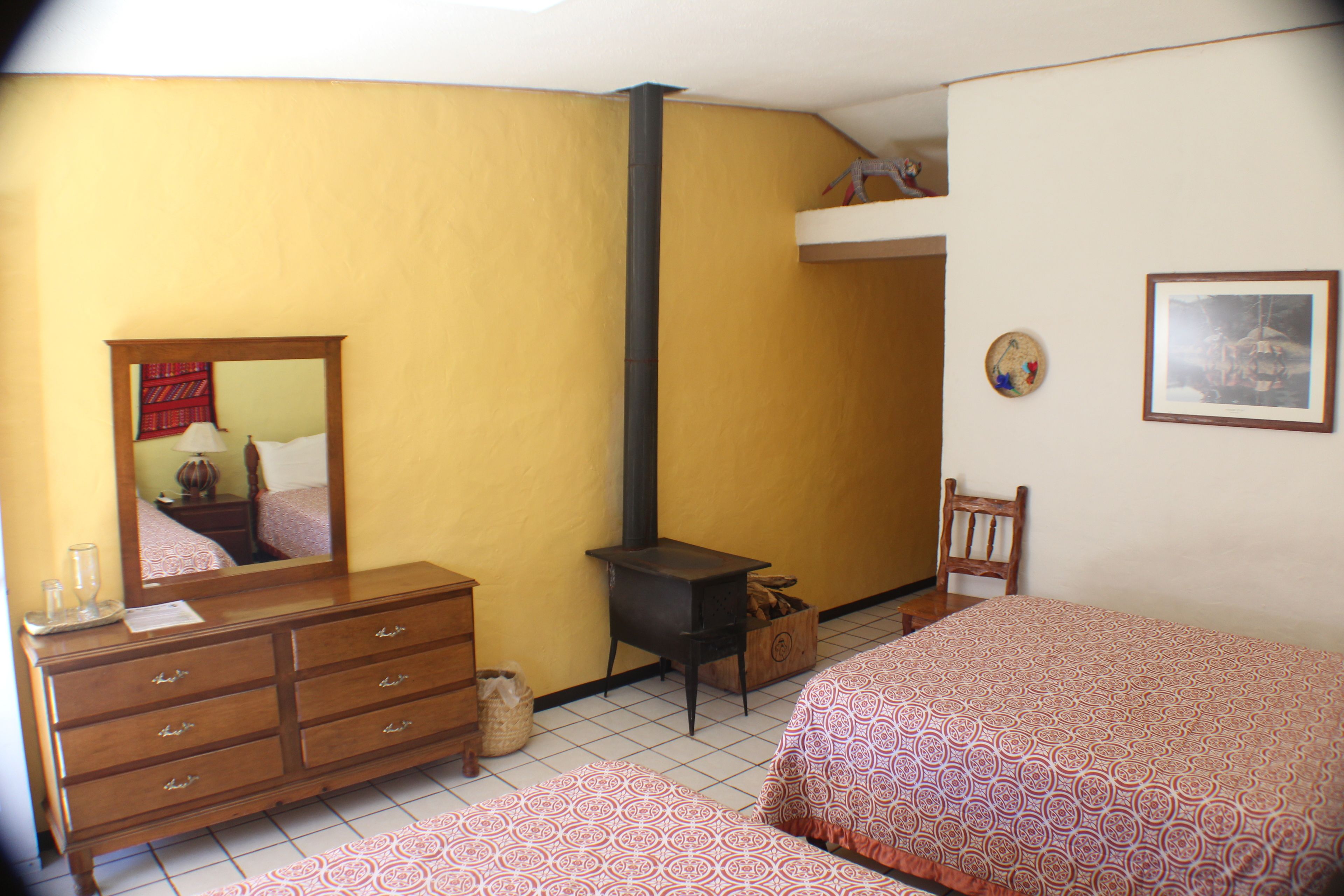 A bedroom with a wooden dresser, a bed with patterned bedding, a small wooden cabinet, a heater, and a mirror.