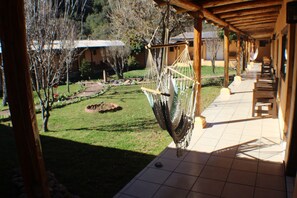 A hammock hanging from a wooden structure in a covered outdoor area with a tiled floor and a view of a grassy yard and buildings in the background.