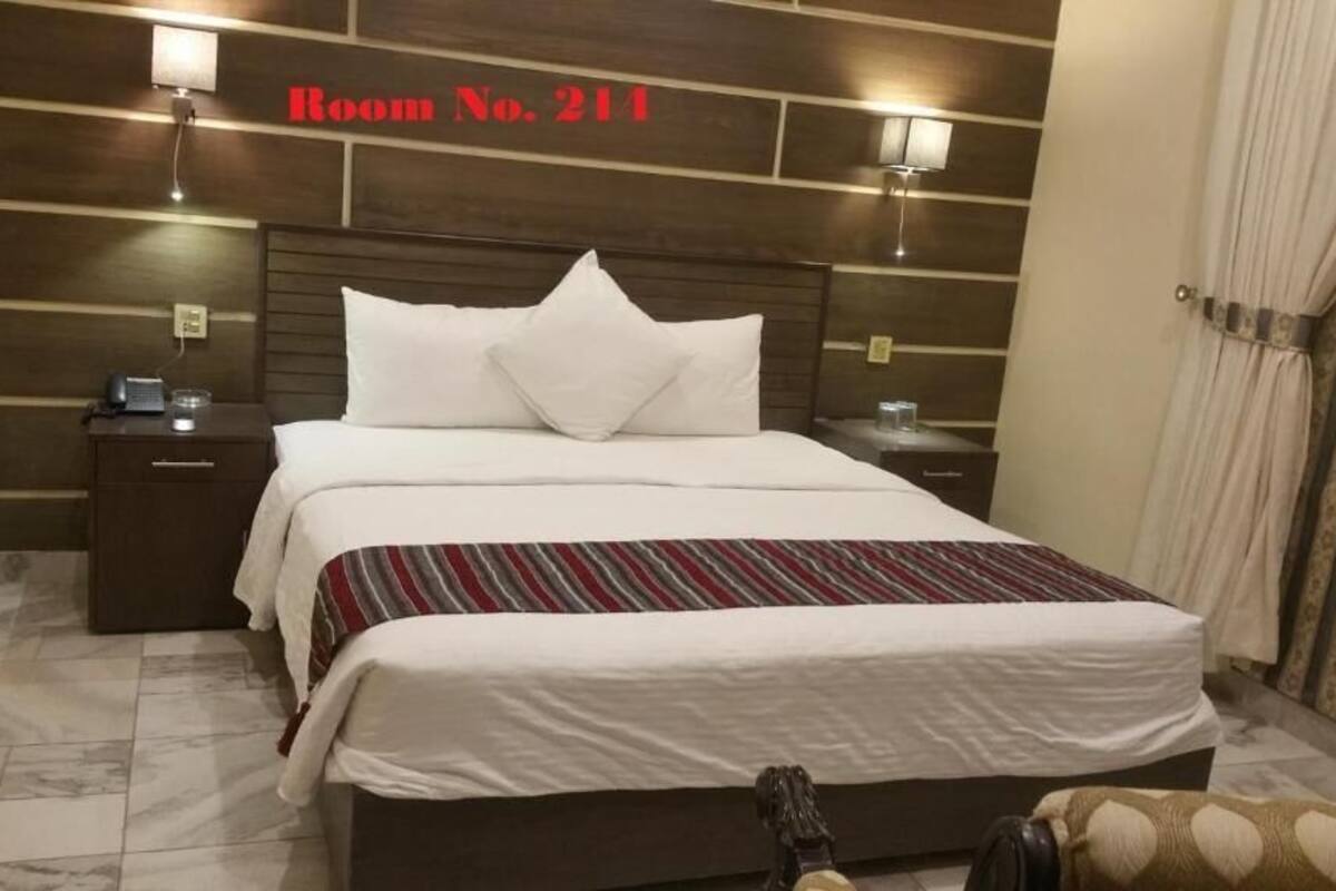 Habitación de hotel con una cama grande, cabecera con diseño de panel de madera, mesita de noche con lámpara y un cartel en la pared que indica el número de habitación.